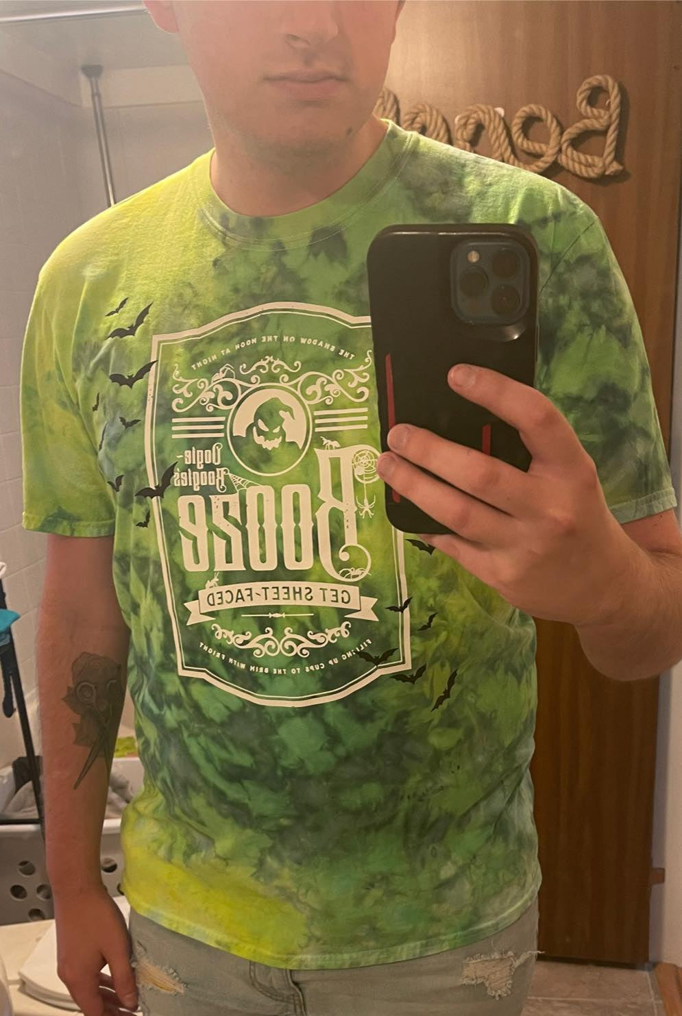 Person wearing a green t-shirt with a graphic design, taking a mirror selfie.